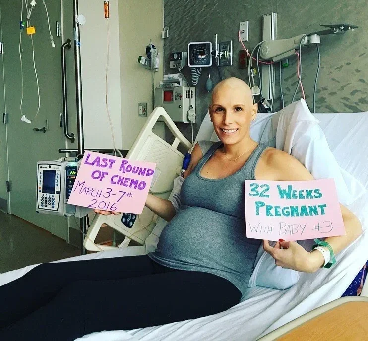 Pregnant woman in a hospital bed holding signs indicating her last round of chemo and her pregnancy at 32 weeks with her third baby.