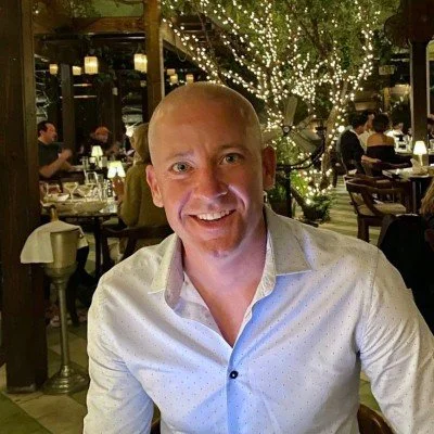 A smiling man in a white shirt sitting at a restaurant with string lights and trees in the background.