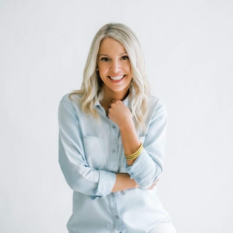 A smiling woman with blonde hair, wearing a light blue shirt and gold bangles, posing against a plain white background.