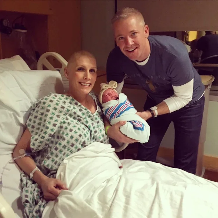 A woman in a hospital bed holding a newborn wrapped in a striped blanket, smiling at the camera. A man is standing next to her, leaning in and smiling, holding the baby's head. The woman appears to be in a hospital gown, and medical tubing is visible on her arm.