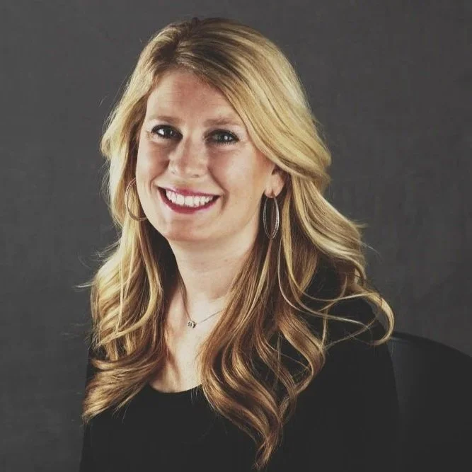 Portrait of a smiling woman with blonde wavy hair wearing black clothing and hoop earrings against a dark background.