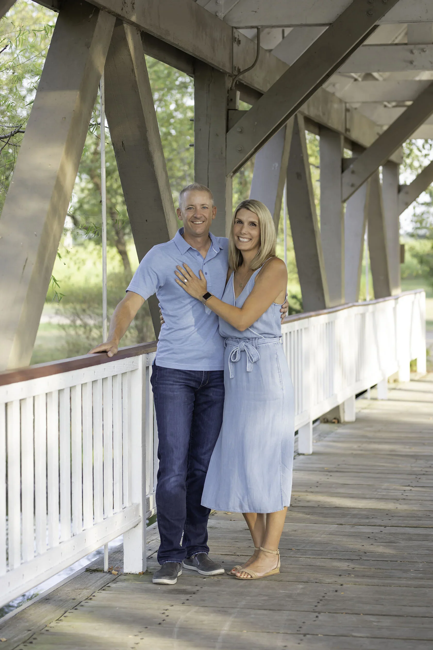 A smiling couple standing on a wooden bridge, embracing each other, with green trees in the background.