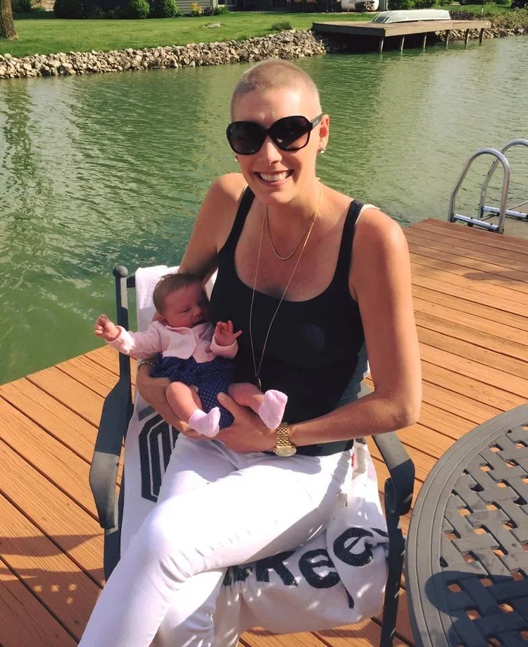 A woman with short hair, sunglasses, and a black tank top sitting on a wooden dock, holding a small baby dressed in pink and blue, near a body of green water with a dock ladder in the background.