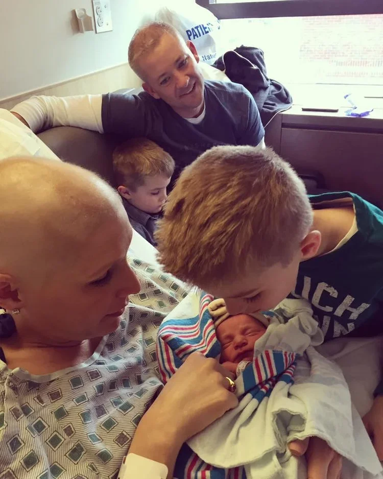 A woman with a shaved head lying in a hospital bed holding a newborn wrapped in a blanket, while a young boy with blond hair kisses the baby's head, and an older man, possibly in a hospital gown, sits nearby in a hospital room.