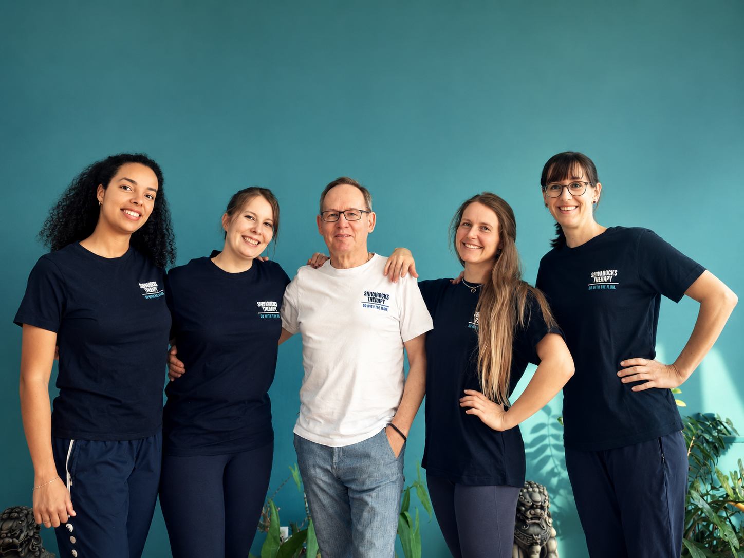 Fünf Menschen stehen zusammen vor einer blauen Wand und lächeln, die meisten tragen schwarze T-Shirts mit weißem Text.