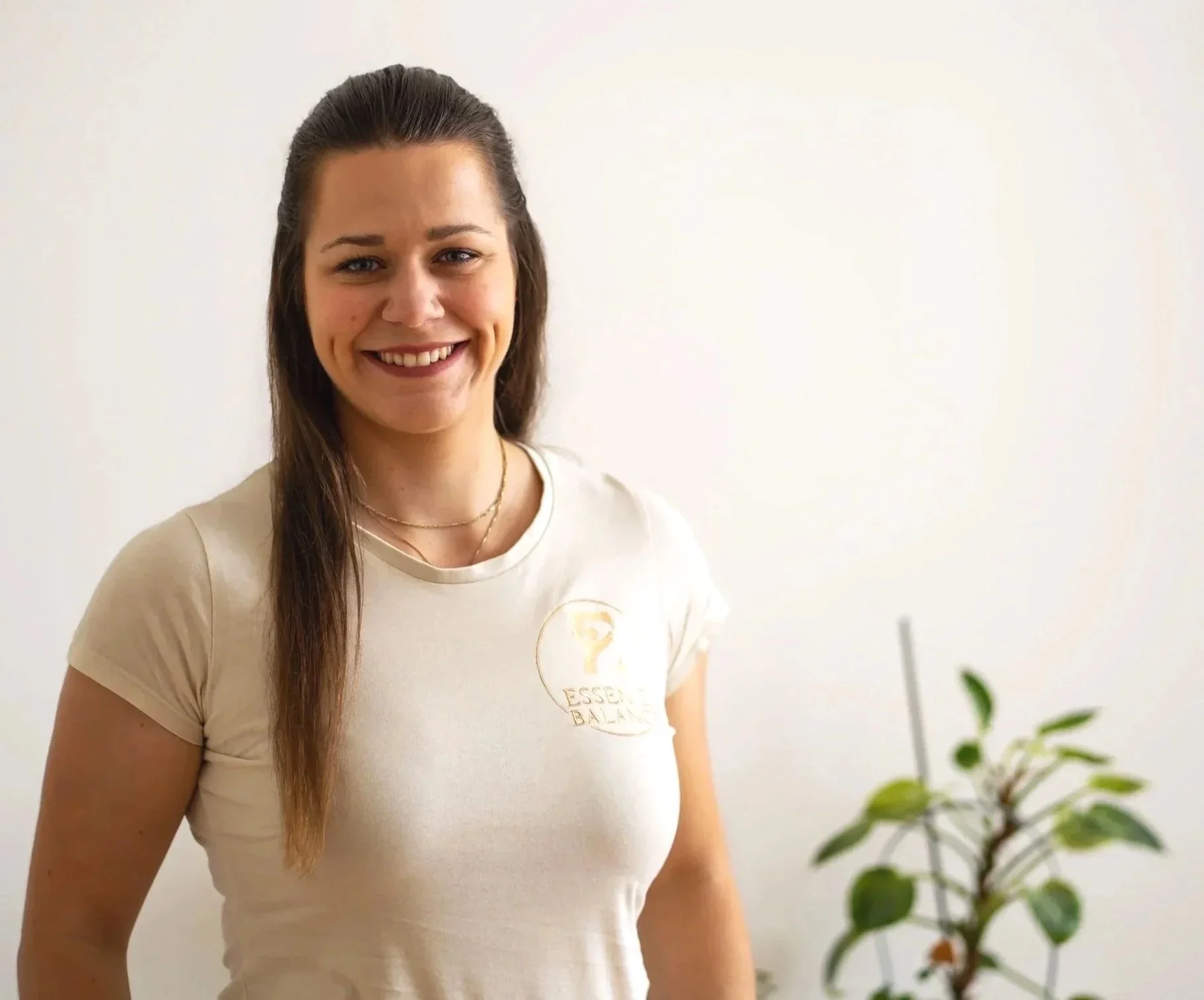 Junge Frau mit braunen Haaren, die an einem weißen Wand lächelt, trägt ein weißes T-Shirt mit einem Logo auf der Brust und steht neben einer grünen Pflanze.