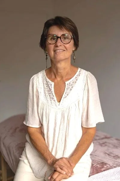 Célina Seignard, femme souriante avec des lunettes, portant un ensemble blanc, dans son cabinet de réflexologie, sophrologie à Vannes