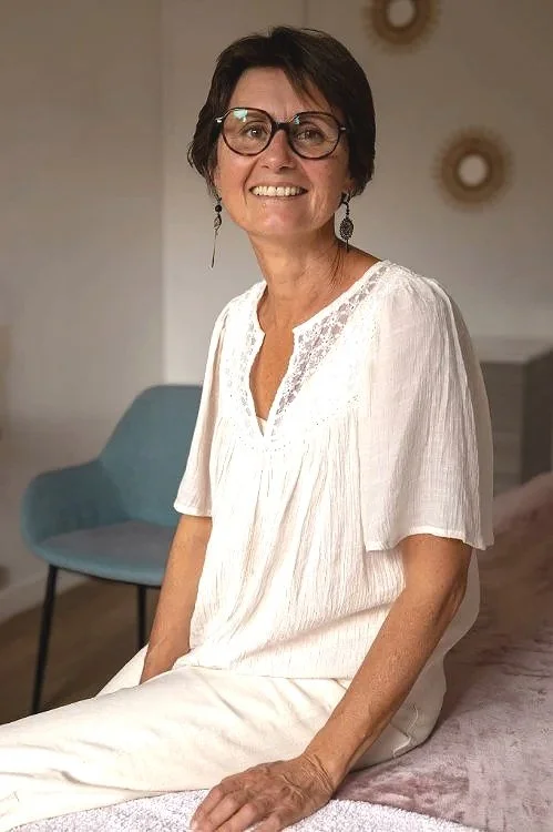 Célina Seignard, femme souriante avec des lunettes, portant un ensemble blanc, dans son cabinet de réflexologie, sophrologie à Vannes