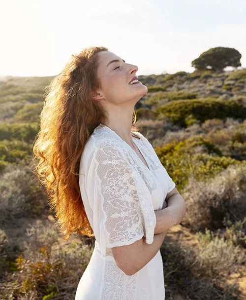 Une femme rieuse avec les yeux fermés, cheveux roux bouclés, portant une robe blanche en dentelle, dans un paysage en plein air avec des buissons, un arbre et le ciel au coucher du soleil.