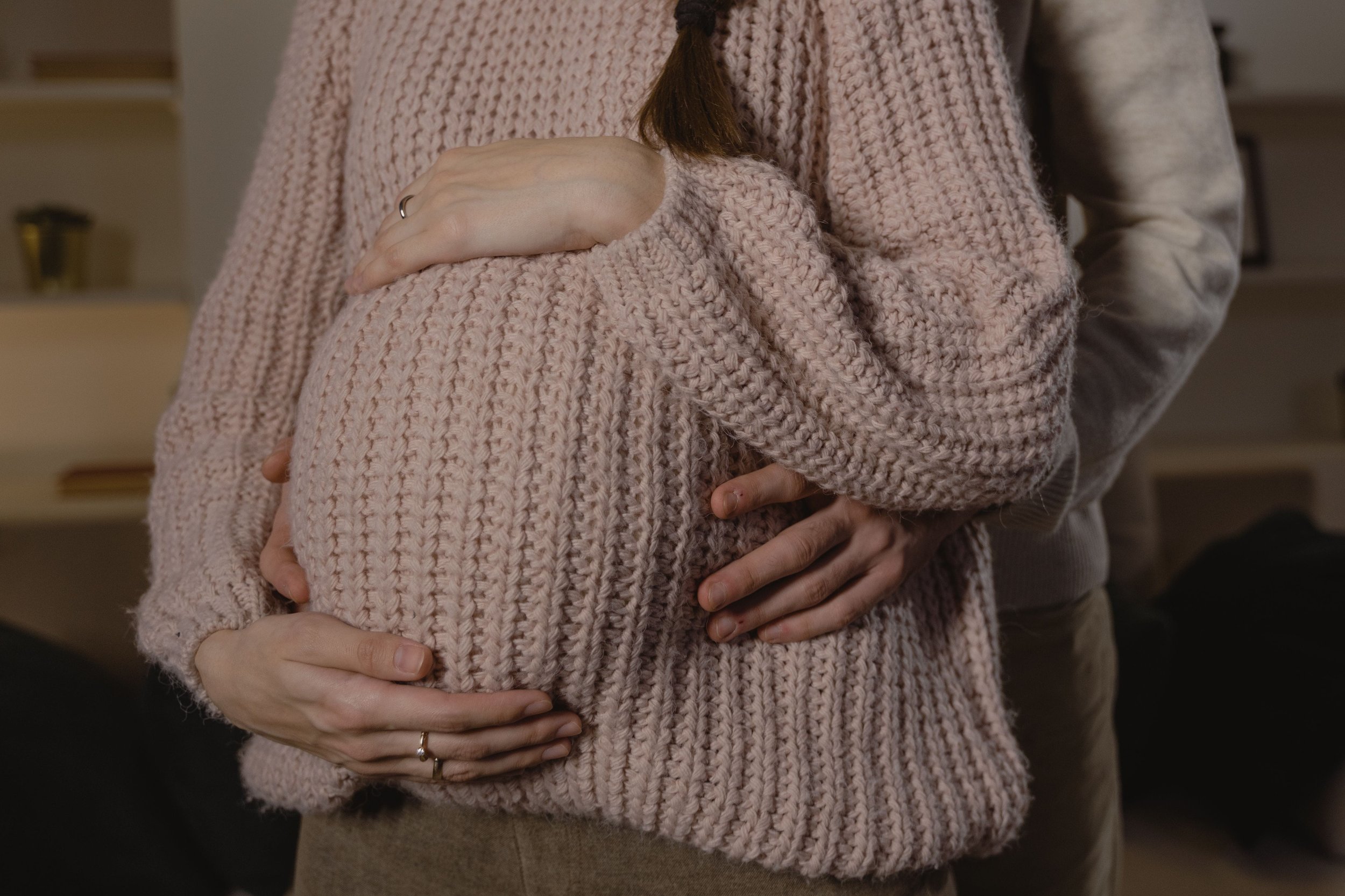 People hugging each other, one wearing a pink knitted sweater, the other holding their hands on the person's stomach.