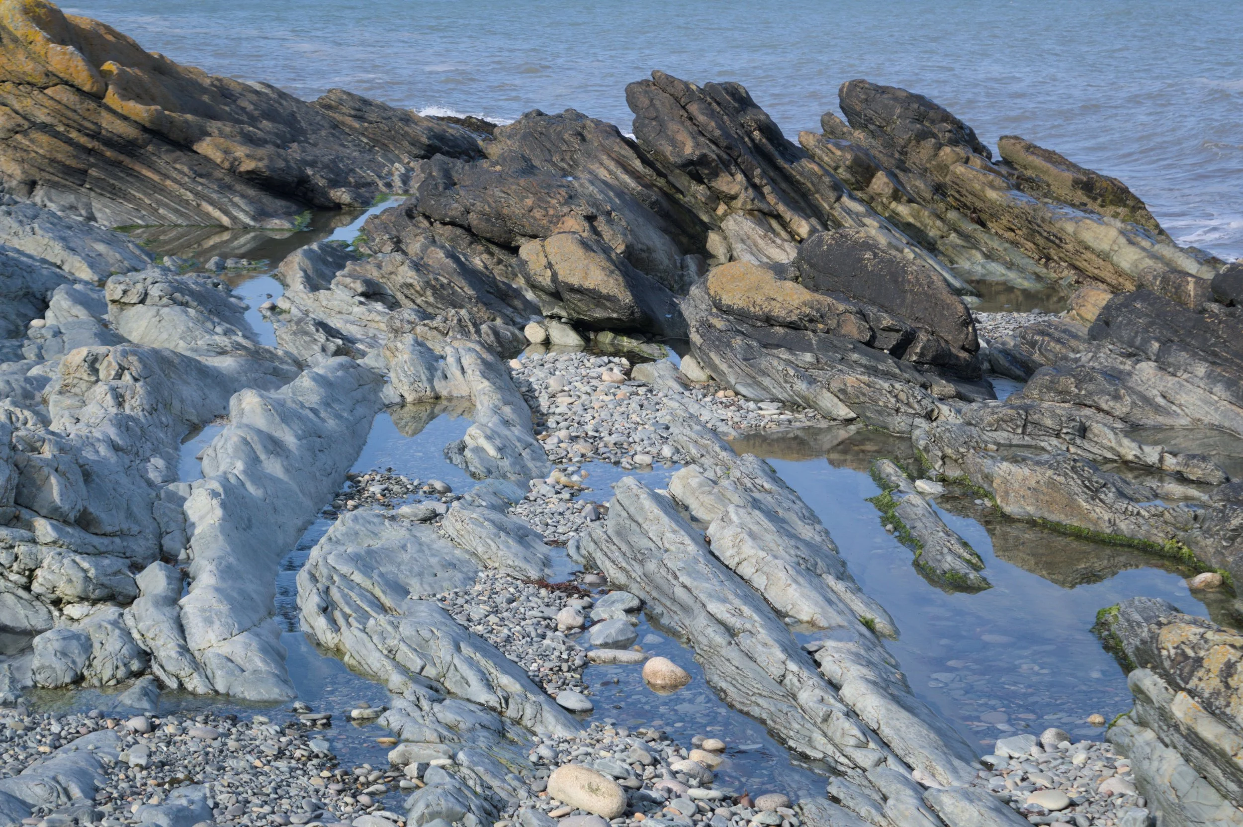 Coastal scene with large, layered rocks and boulders along a shoreline, with water in the background and some tide pools formed among the rocks.