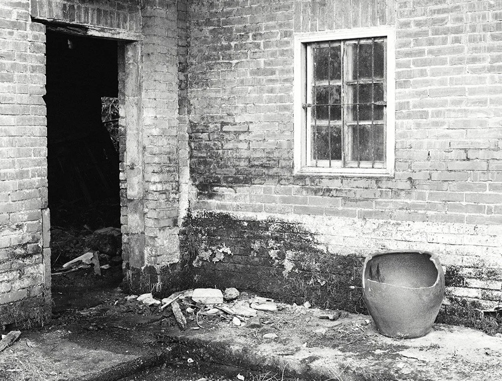 Black and white photo of a brick house with a window, doorway, and a large ceramic pot on the ground.