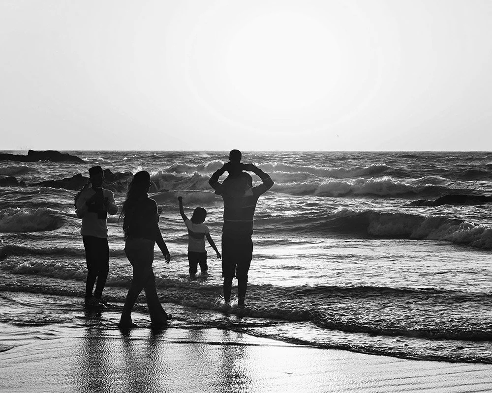 Family enjoying time at the beach, with four people silhouetted against the ocean waves, one child sitting on an adult's shoulders, late afternoon or early evening lighting.
