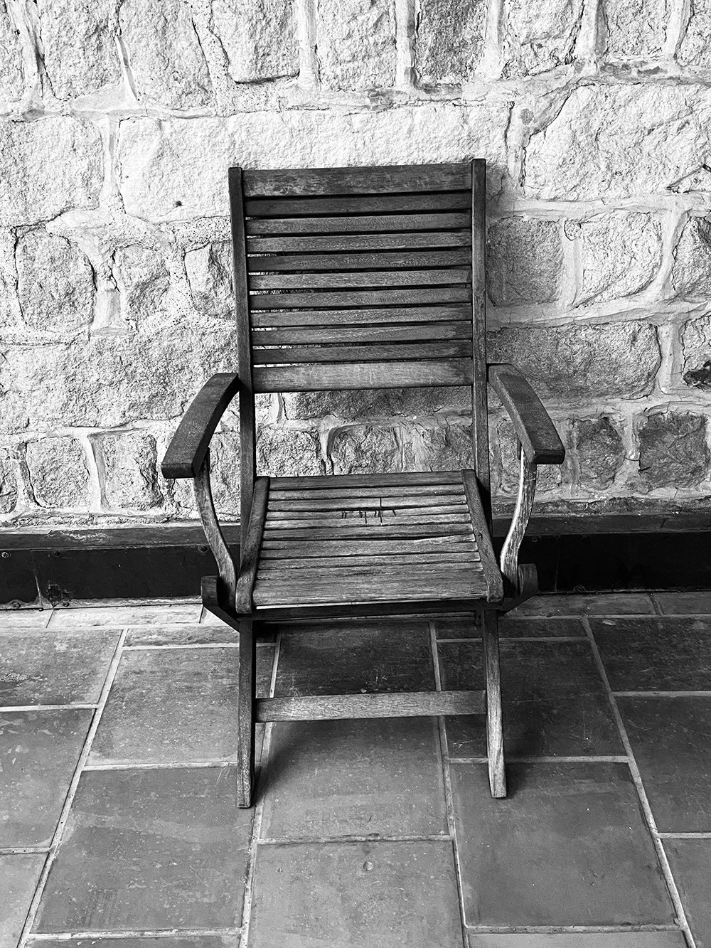 A wooden chair with armrests is placed against a textured stone wall, on a tiled floor.