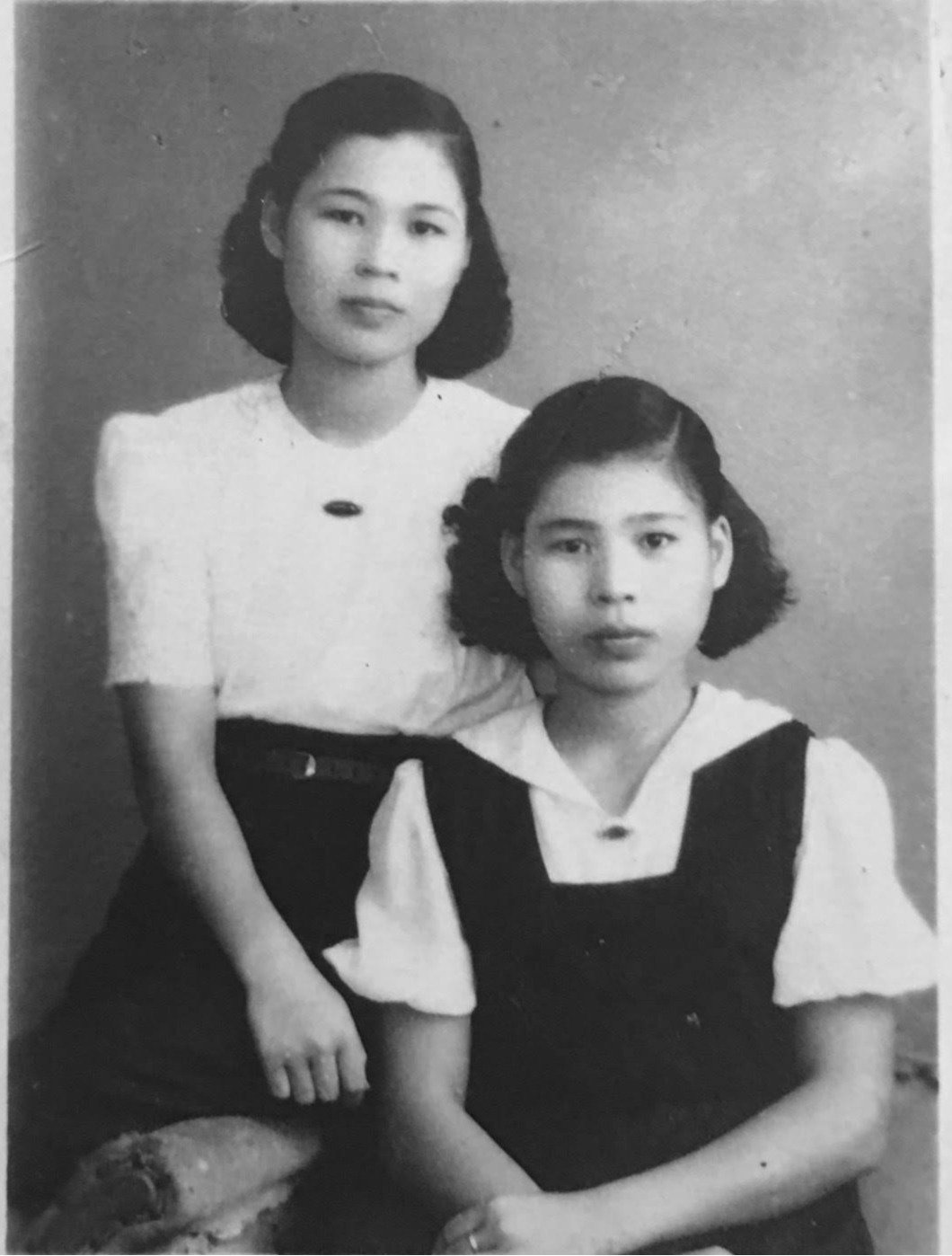 Black and white photo of two young girls with shoulder-length hair, one standing behind the other, both wearing dresses with collared shirts. They are posing against a plain wall.