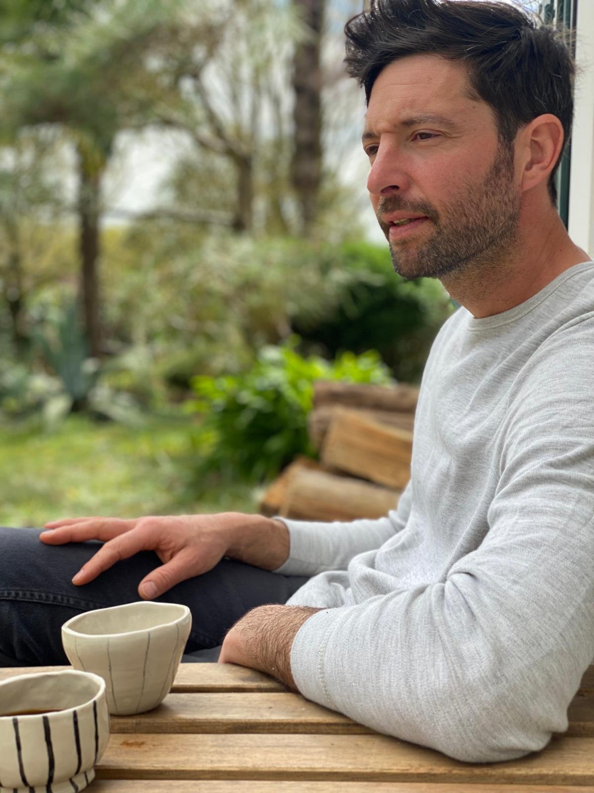 Un homme assis à une table en bois à l'extérieur, avec des tasses à café ou thé devant lui, entouré de nature avec des arbres en arrière-plan.