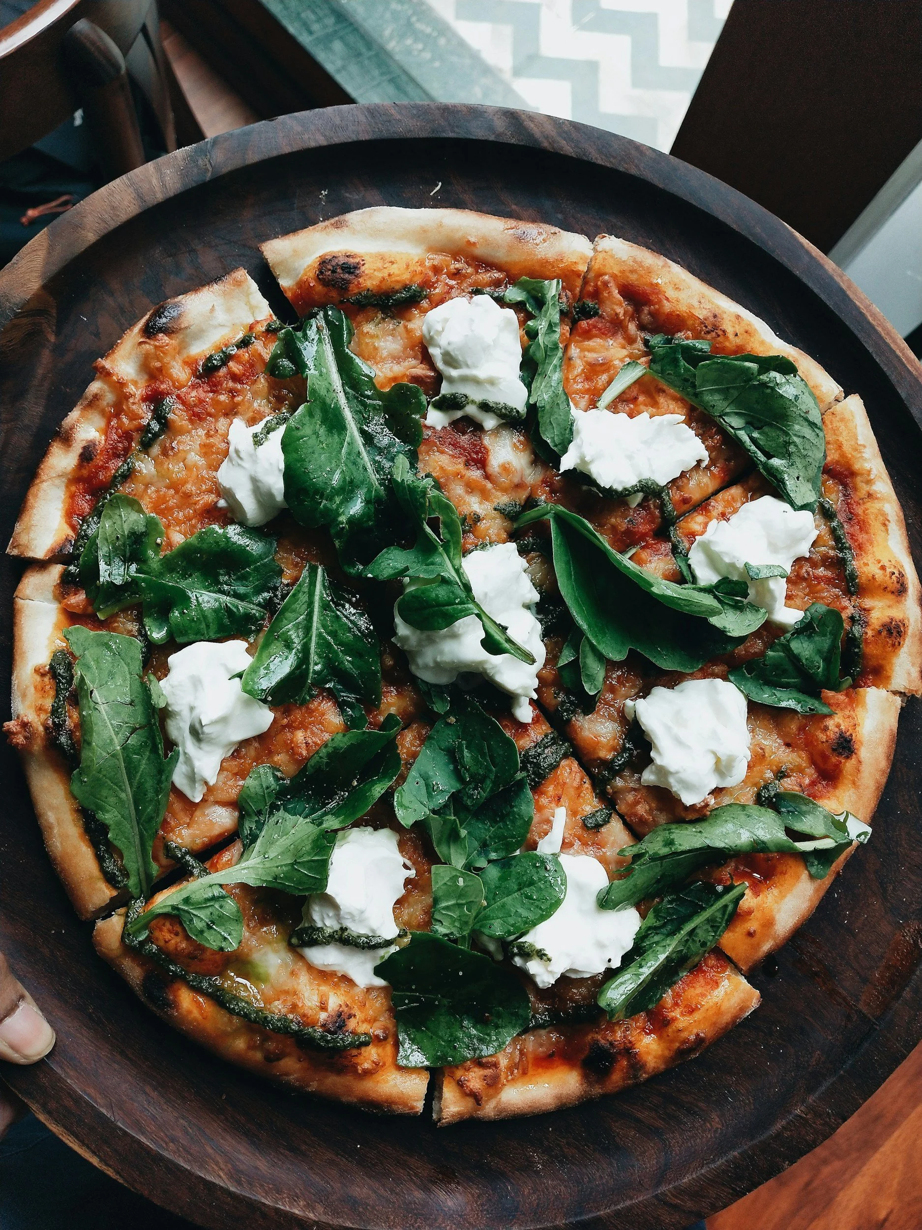 An oven fired pizza with a thin crust topped with ricotta cheese, fresh spinach, grilled zucchini, and tomato sauce, served on a wooden pizza tray in the Hamptons.