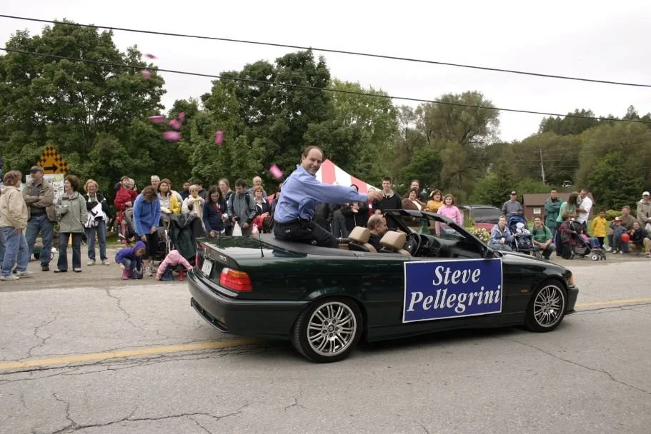 A dark green convertible car with Steve Pellegrini sitting on the back seat, waving pink objects. The car has a sign that says 'Steve Pellegrini'. In the background, a crowd of people is gathered, some children and adults, watching the parade.