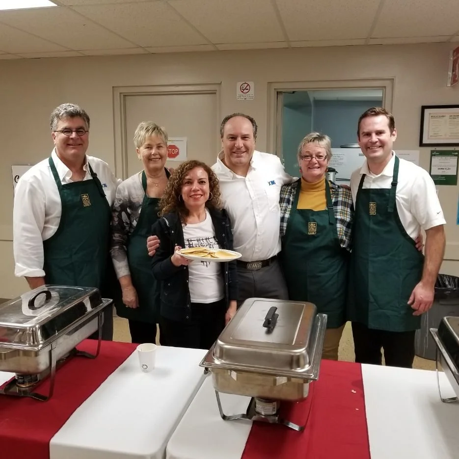 Group of six people wearing aprons, standing behind a table with serving trays in a community room or cafeteria, celebrating or participating in a food event.