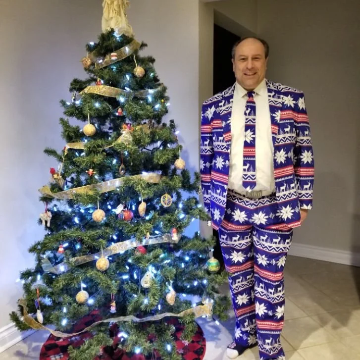 Mayor Steve Pellegrini in a blue and white holiday patterned suit standing next to a decorated Christmas tree with lights and ornaments.