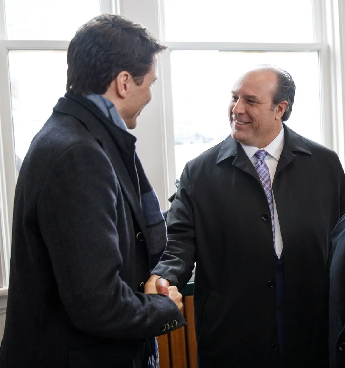 Two men in dark coats shaking hands and smiling at each other indoors near a window.