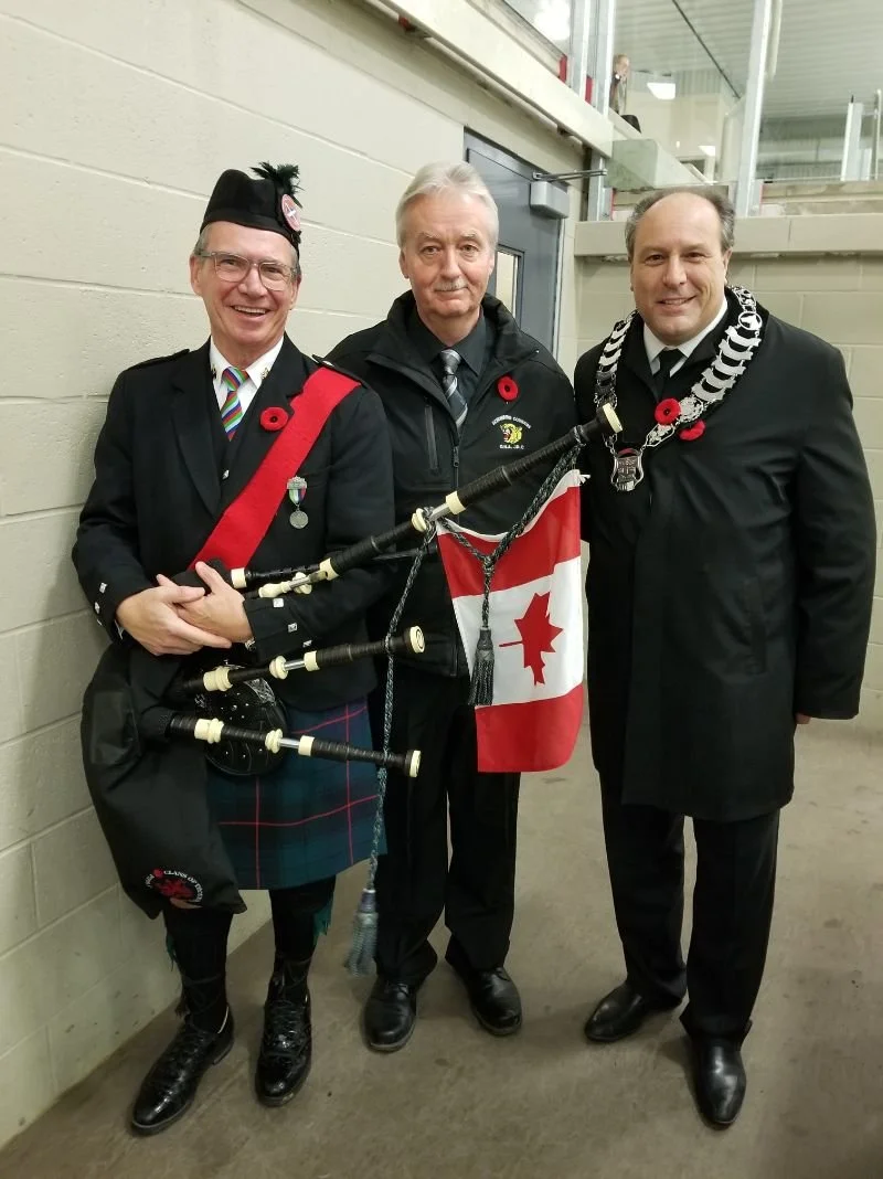 Three men dressed in formal and traditional attire, one holding a bagpipe with the Canadian flag, standing in a hallway.