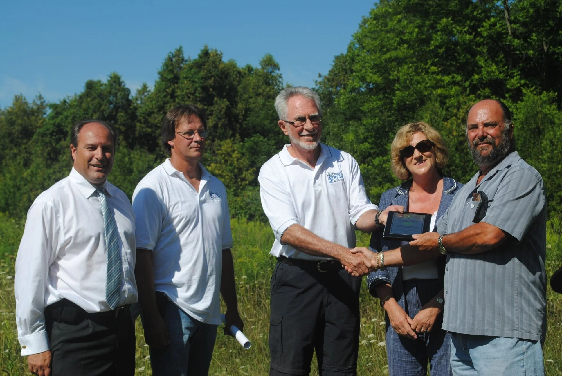 A group of five people standing outdoors, with a man and a woman shaking hands and exchanging a tablet device. They are in a green field with trees and a blue sky in the background, dressed in semi-formal attire.