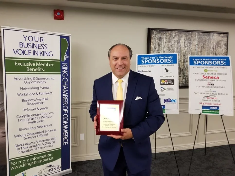 	
Mayor Steve Pellegrini holding an award plaque, standing in front of promotional signs for the King Chamber of Commerce at an event.