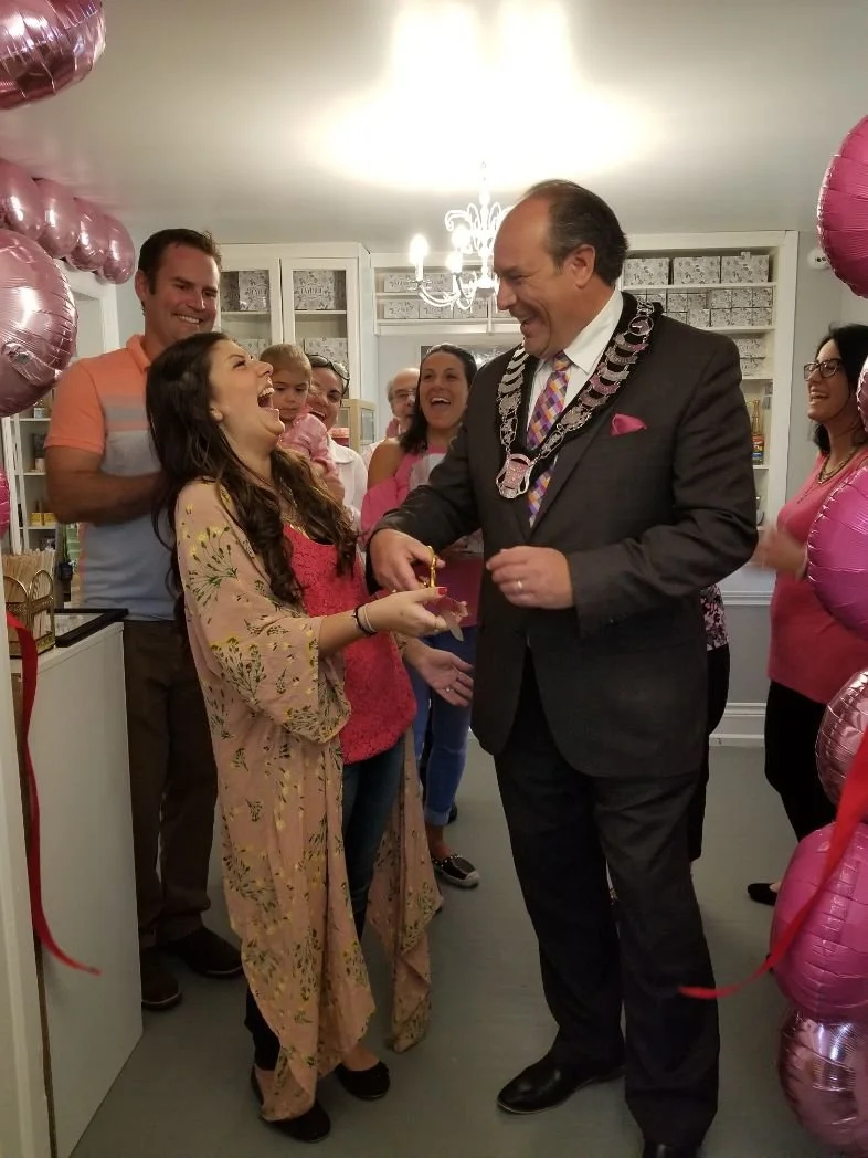 A woman receives a necklace from Mayor Steve Pellegrini wearing a formal suit and chain, family and friends smile and laugh around them at a celebration with pink balloons.