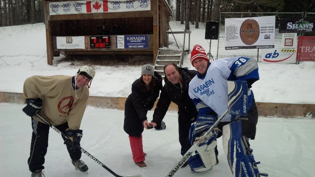 Four people on an outdoor ice rink with snow in the background, posing for a photo. One person is wearing hockey gear, including pads and a helmet, and holding a hockey stick; another is in casual winter clothing with red boots; two women are in dark