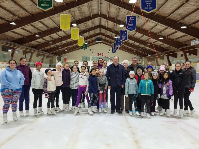 A group of ice skaters with Mayor Steve Pellegrini inside an ice rink, with banners hanging from the wooden ceiling.