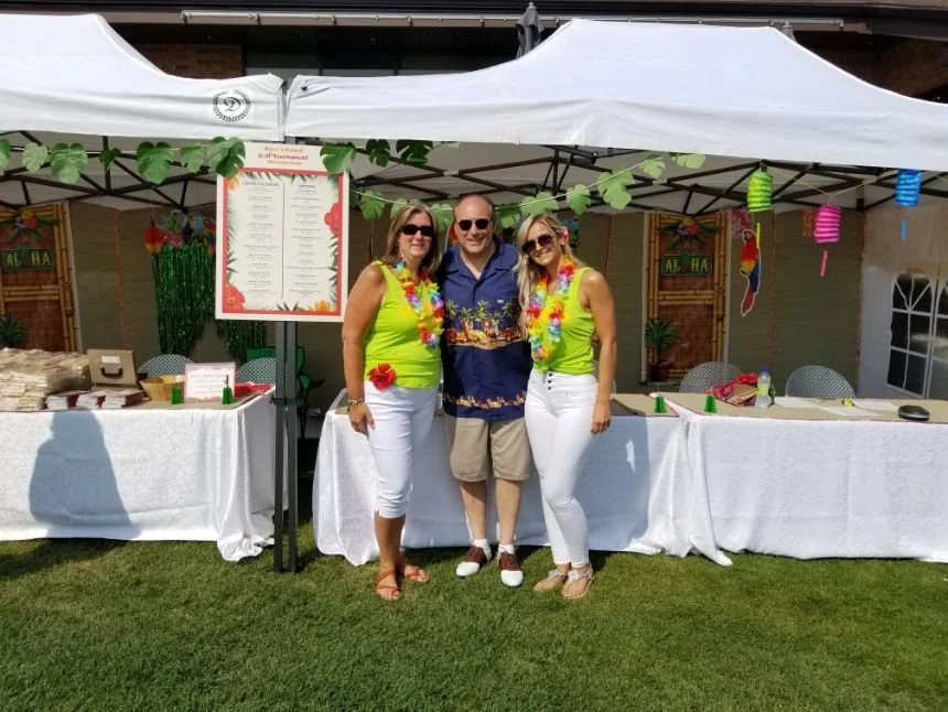 	
Mayor Steve Pellegrini posing outside at a tropical-themed party under a white canopy with green leaves, colorful decorations, and a menu board on display.