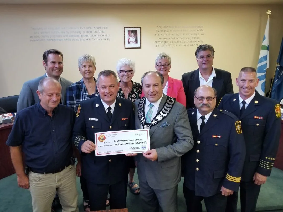 Group of people with Mayor Steve Pellegrini celebrating with five individuals in firefighter uniforms holding a large check for five thousand dollars, surrounded by others in business attire, in a room with a framed picture and flags.