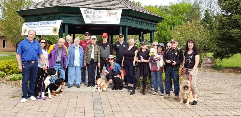 Group of people with Mayor Steve Pellegrini and their dogs standing in front of a shelter in a park, with trees and a lawn in the background.