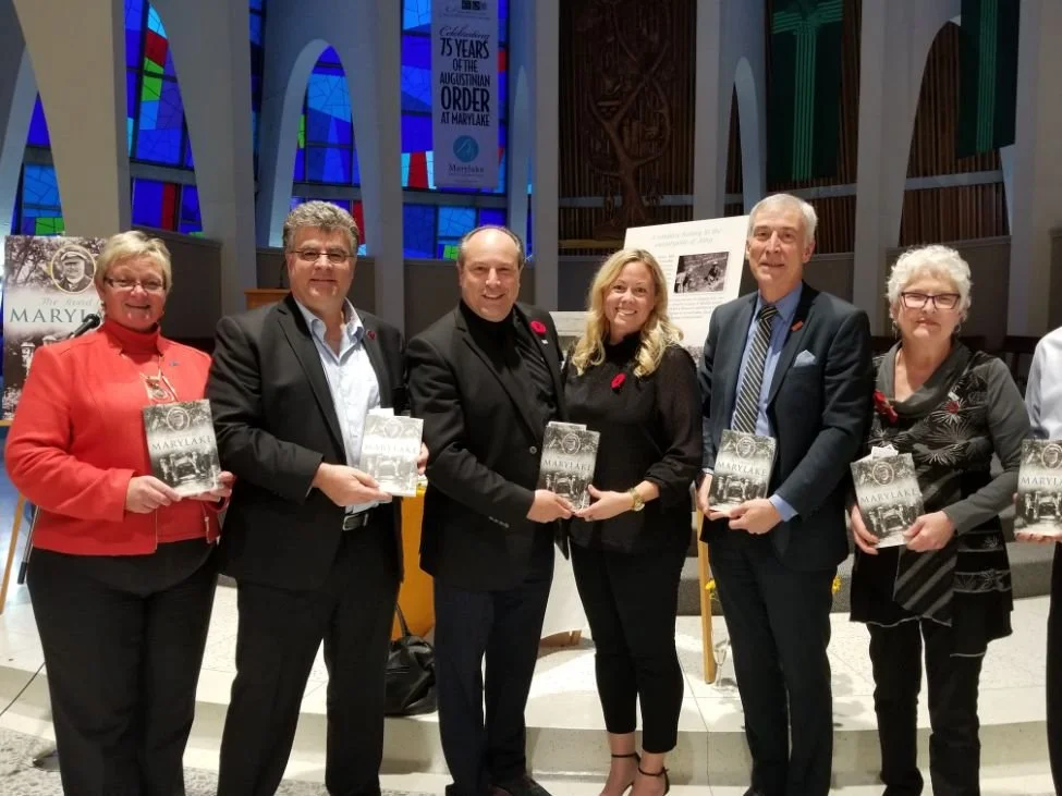 	
Mayor Steve Pellegrini standing in a row inside a church, holding books titled 'Mary Lake'. The church features stained glass windows and informational displays in the background.