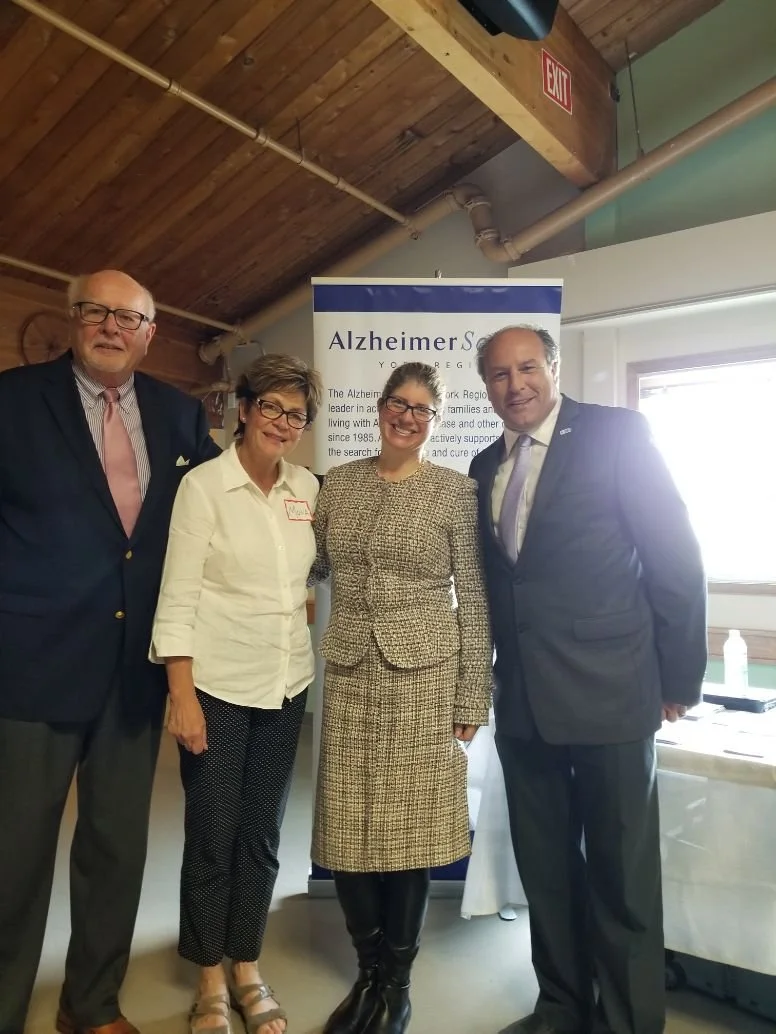 Four people pose in front of an Alzheimer's Society banner indoors with wooden ceiling and ductwork visible, three are smiling and dressed professionally, one woman with glasses and two men in suits.