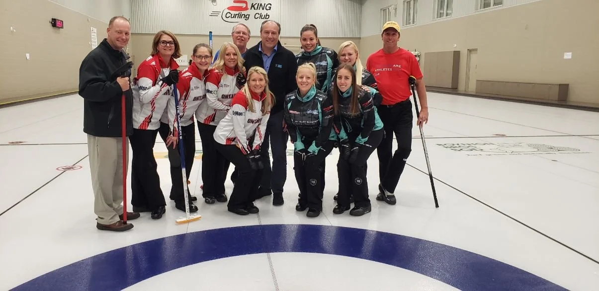 Group of people, mostly women, standing on an indoor curling rink, smiling at the camera, some holding curling brooms and wearing curling uniforms, with a sign that reads 'King Curling Club' in the background.