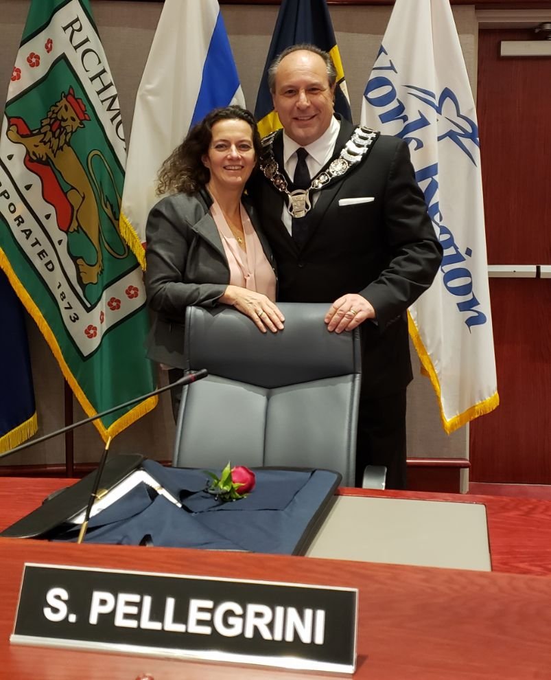 A man and a woman standing together at a formal event, with flags behind them and a sign reading 'S. Pellegrini' on the table in front. The man is wearing a suit with a ceremonial chain, and the woman is dressed in formal attire.