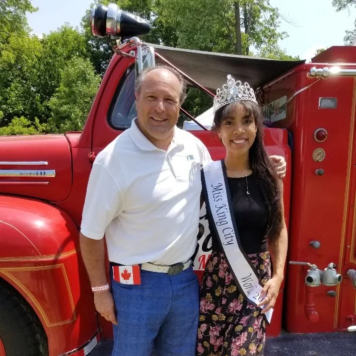 A young woman wearing a sash and tiara, standing next to a man in front of a red fire truck, outdoors with trees in the background.