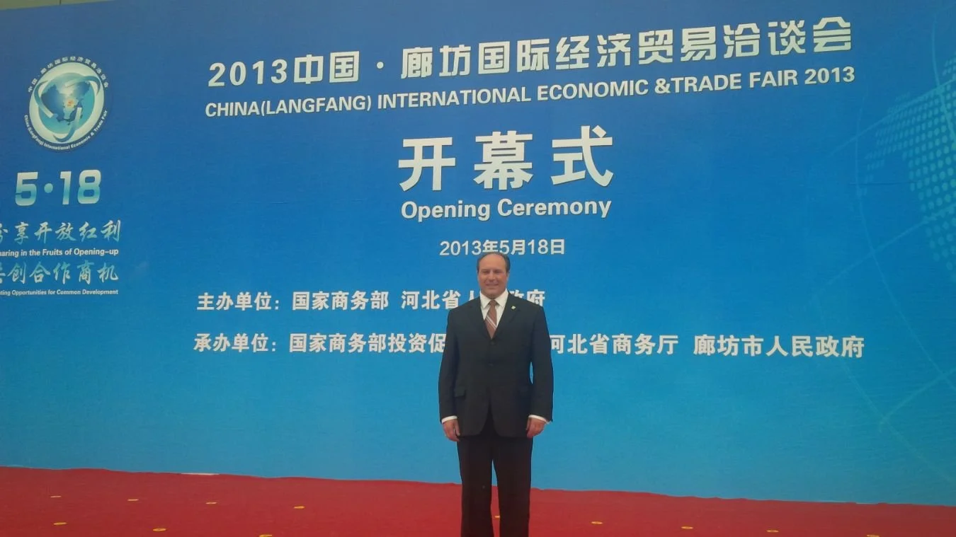 Mayor Steve Pellegrini standing in front of a large blue backdrop at the opening ceremony of the 2013 China (Langfang) International Economic & Trade Fair, held on May 18, 2013.