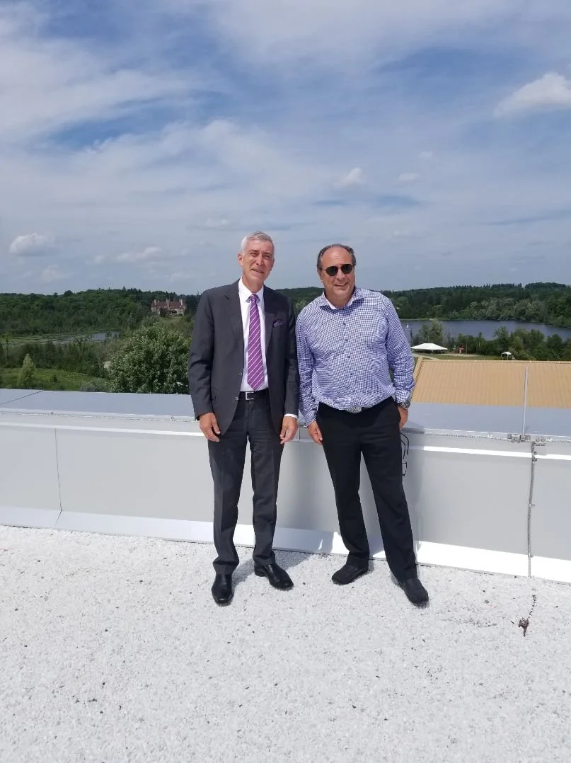 Mayor Steve Pellegrini standing on a rooftop against a scenic background of trees, a lake, and a partly cloudy sky.