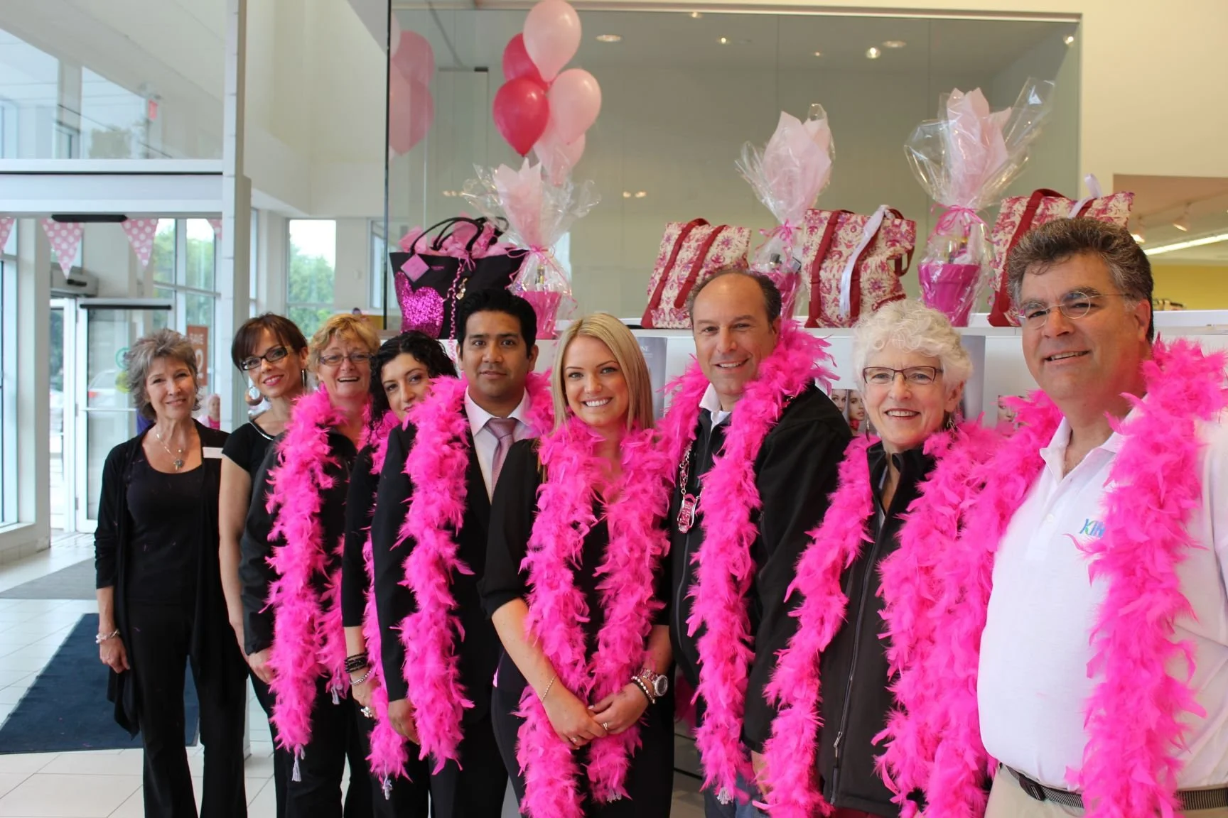 Group of nine people dressed in black and white, wearing pink feather boas, standing indoors with pink balloons and gift bags in the background, celebrating an event.