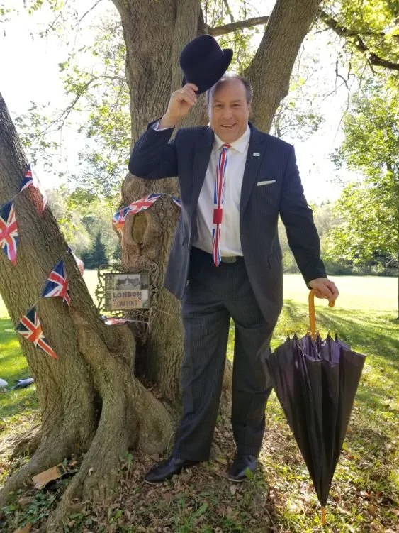 	
Mayor Steve Pellegrini holding a bowler hat, standing outdoors next to a tree decorated with Union Jack flags and a London-themed sign, holding a black umbrella with a yellow handle.