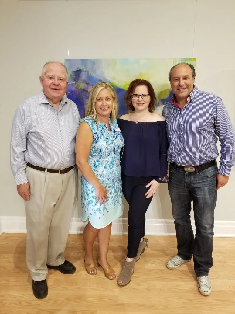 Four adults standing together in a room with a colorful abstract painting behind them, smiling at the camera.