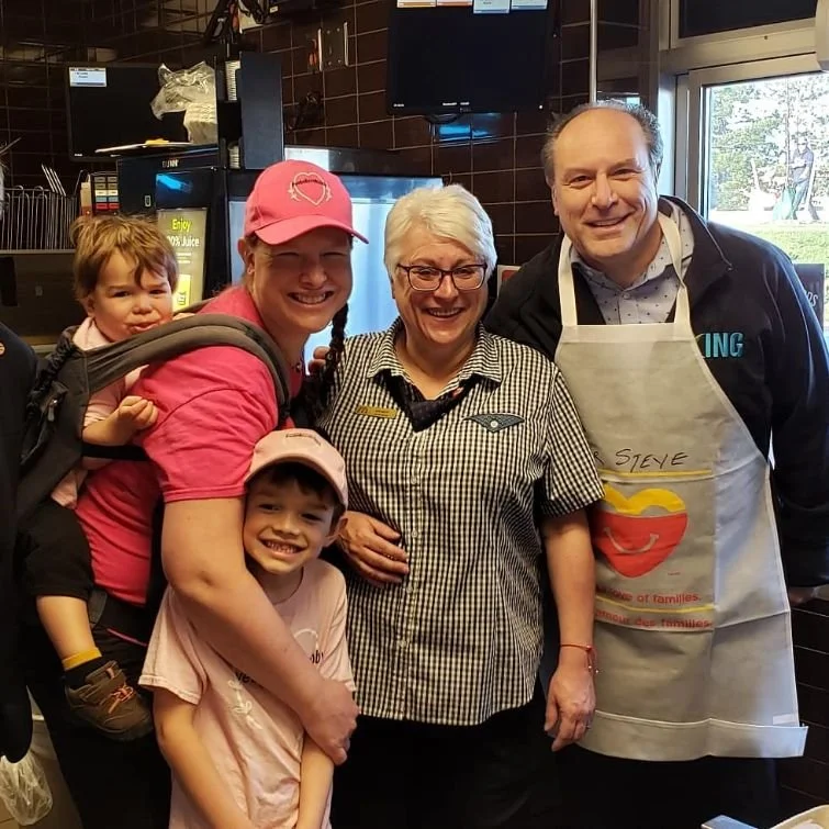 Four people, including two children, a woman, and a man, inside a restaurant. The woman and man are smiling, and the children are posing happily. The man is wearing an apron with writing on it. There are kitchen and digital screens visible in the bac