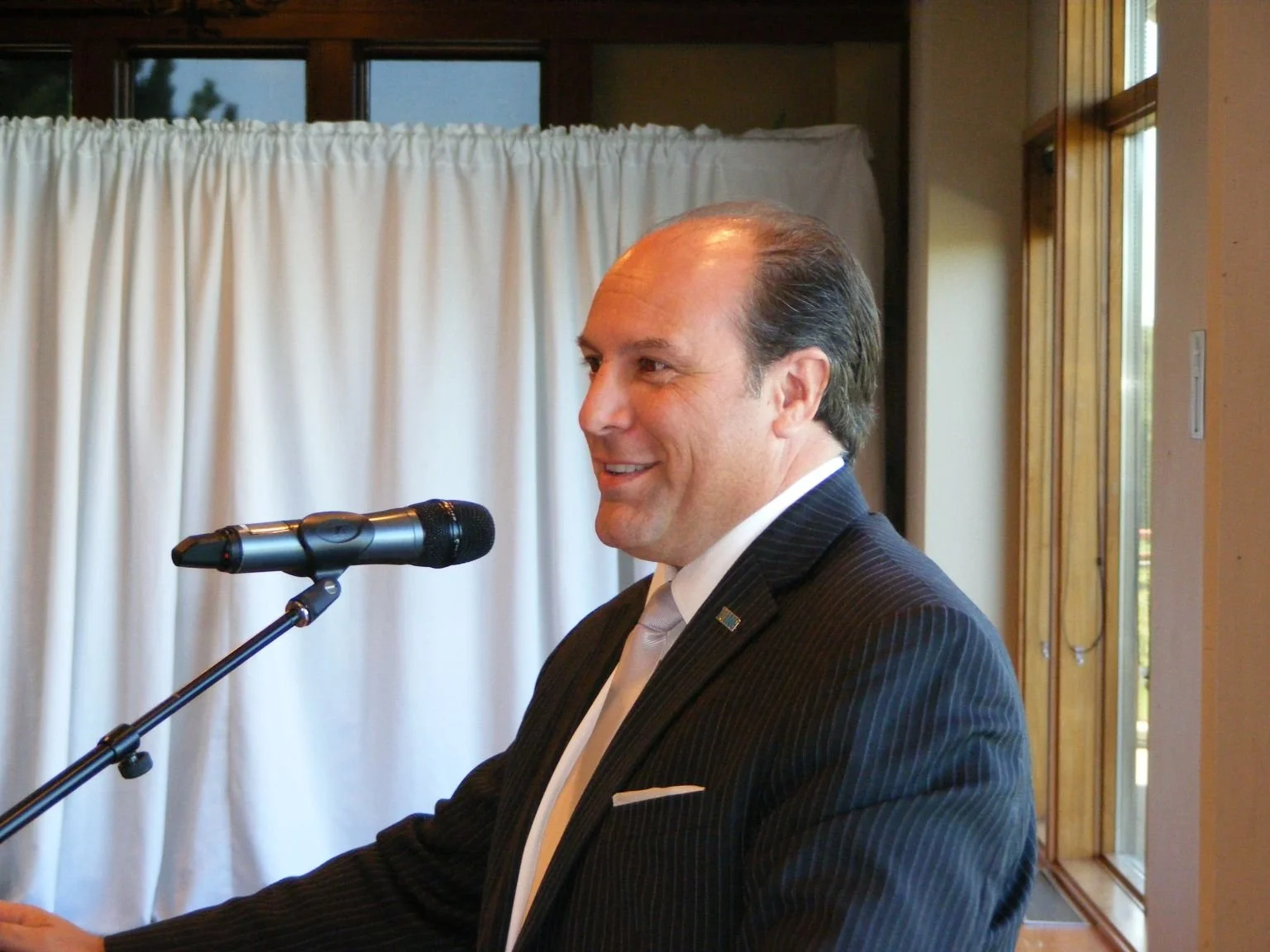 A man in a black suit and white tie standing at a microphone, smiling, with a backdrop of white curtains and wooden windows.