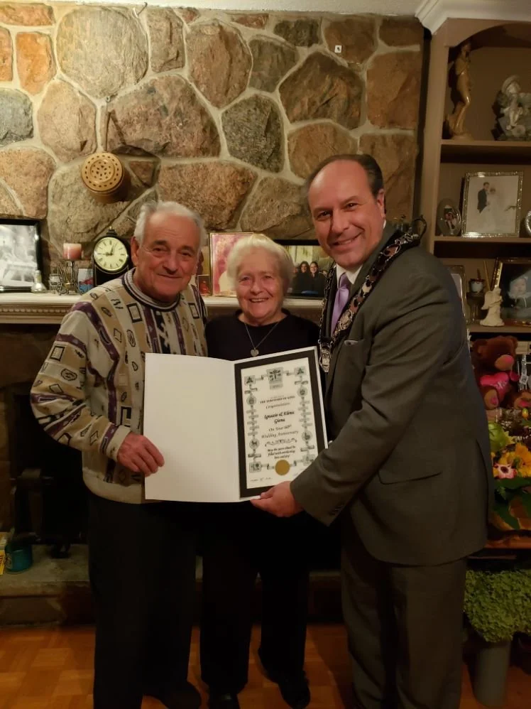 Three people standing in front of a stone fireplace, holding a certificate. An older man with gray hair, an elderly woman with white hair, and a man in a suit smiling at the camera.