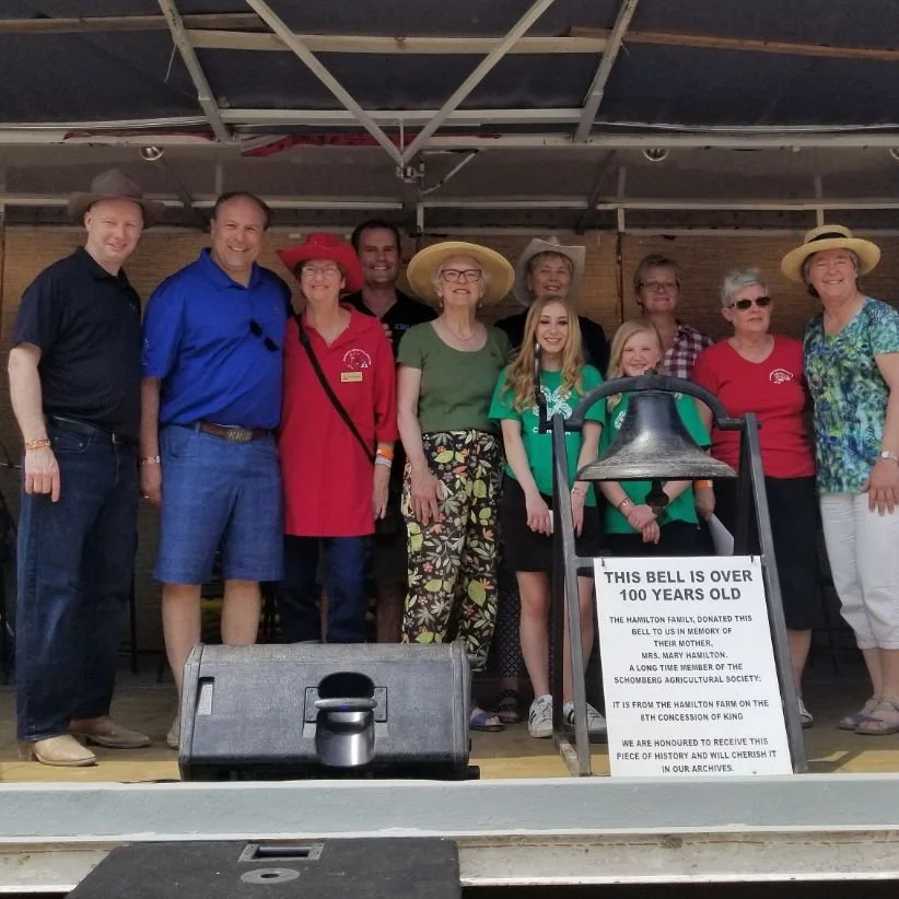 Group of people on a stage, standing behind a sign and a bell, celebrating the bell's 100th anniversary, with a wooden backdrop and a roof overhead.