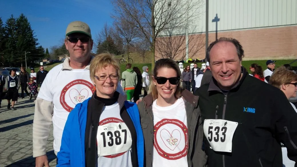 Four people posing outdoors during a race event, with some wearing race bibs and t-shirts with a heart-themed logo, other participants and a building visible in the background.