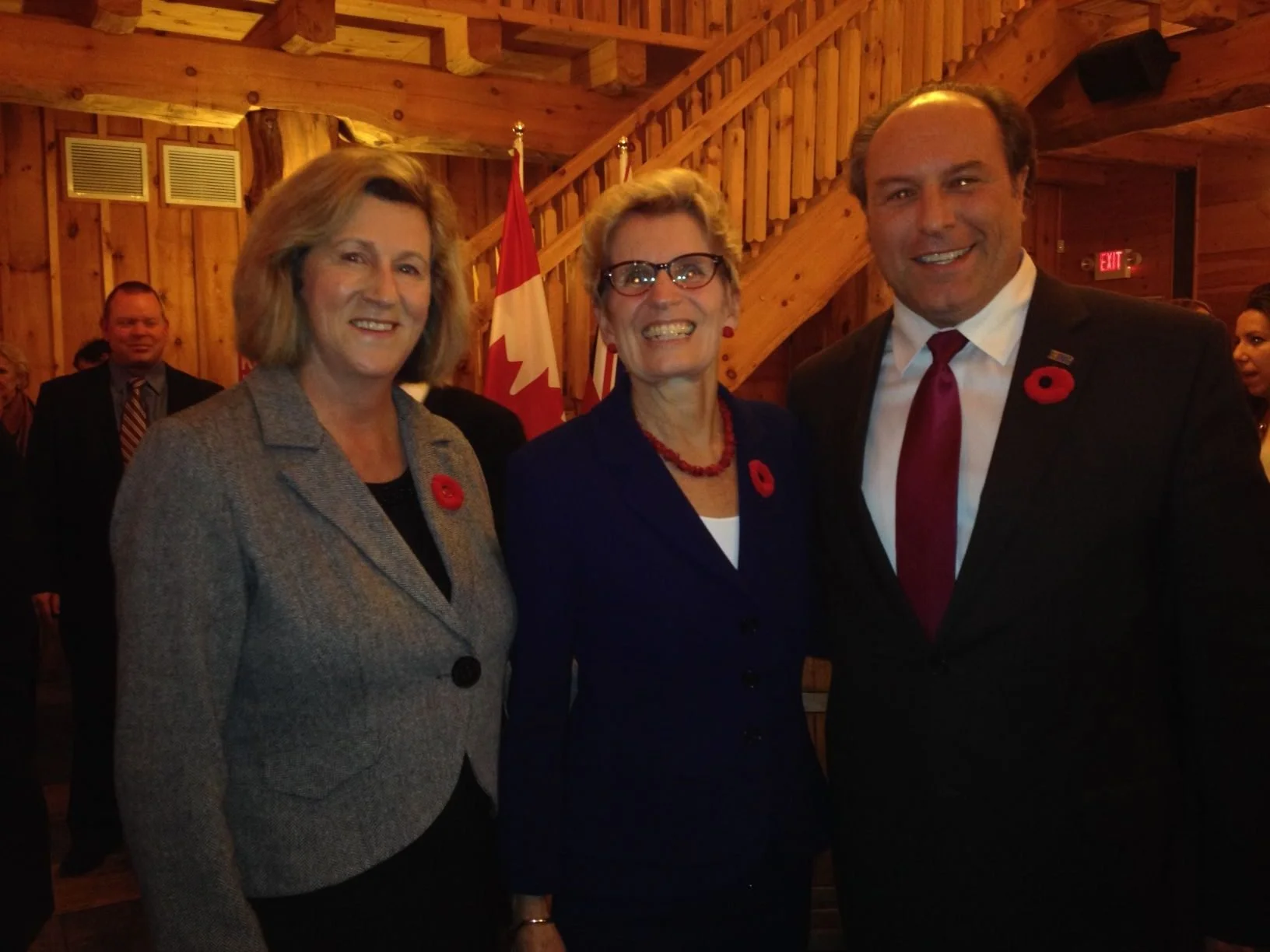 Three smiling individuals, two women and one man, at a formal event with Canadian flags in the background, all wearing poppy pins on their clothing.
